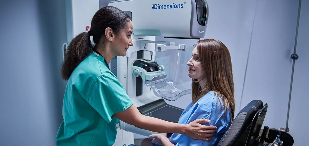 Women sitting in front of a mammography machine with a health care professional at her side.